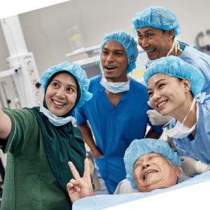 angled photo of smiling surgical team and patient in surgical setting, taking a selfie. Patient is lying down, smiling at the phone camera, making the peace sign. Selfie is being taken by a staff member. Everyone is wearing a blue paper cap.