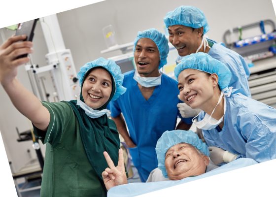 angled photo of smiling surgical team and patient in surgical setting, taking a selfie. Patient is lying down, smiling at the phone camera, making the peace sign. Selfie is being taken by a staff member. Everyone is wearing a blue paper cap.