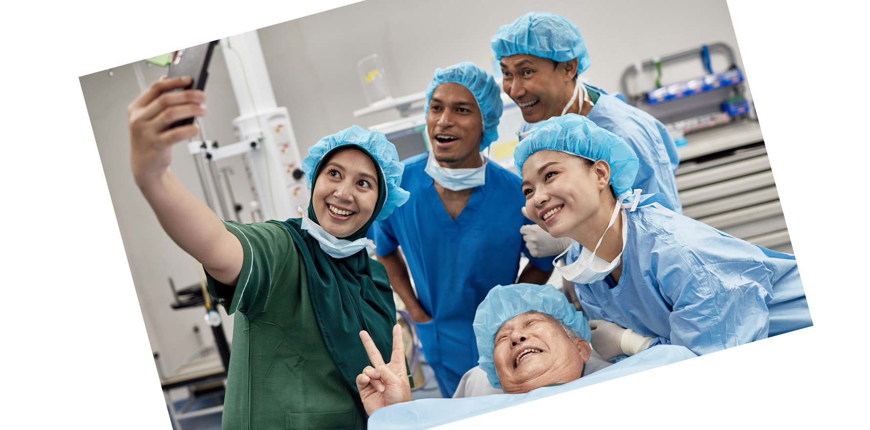 angled photo of smiling surgical team and patient in surgical setting, taking a selfie. Patient is lying down, smiling at the phone camera, making the peace sign. Selfie is being taken by a staff member. Everyone is wearing a blue paper cap.
