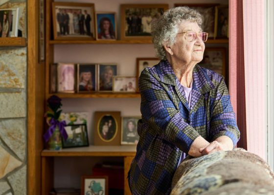 Senior woman standing in front of shelves of family photographs, looking out a window.