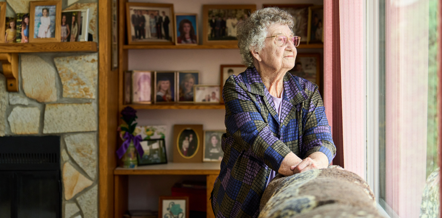 Senior woman standing in front of shelves of family photographs, looking out a window.
