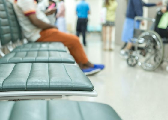 A hospital waiting room, one person sitting at the end of a row or chairs, others in the background.