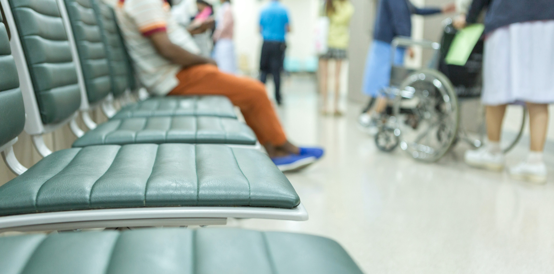 A hospital waiting room, one person sitting at the end of a row or chairs, others in the background.