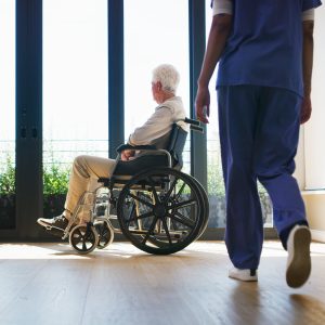 Older person in wheelchair, looking out a window, with a person standing closer to the foreground