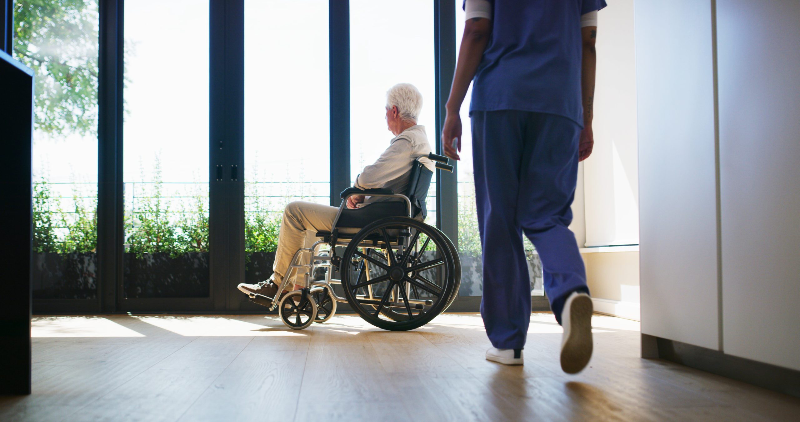 Older person in wheelchair, looking out a window, with a person standing closer to the foreground